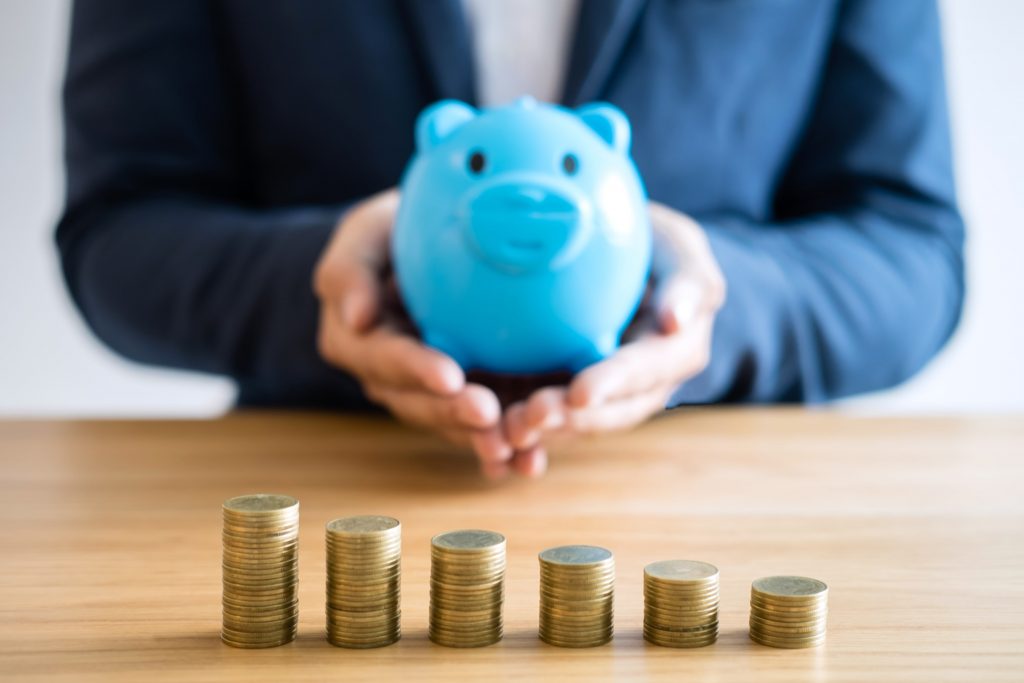 vecteezy_business-person-holding-blue-piggy-bank-at-office-desk-with-stacks-of-coins